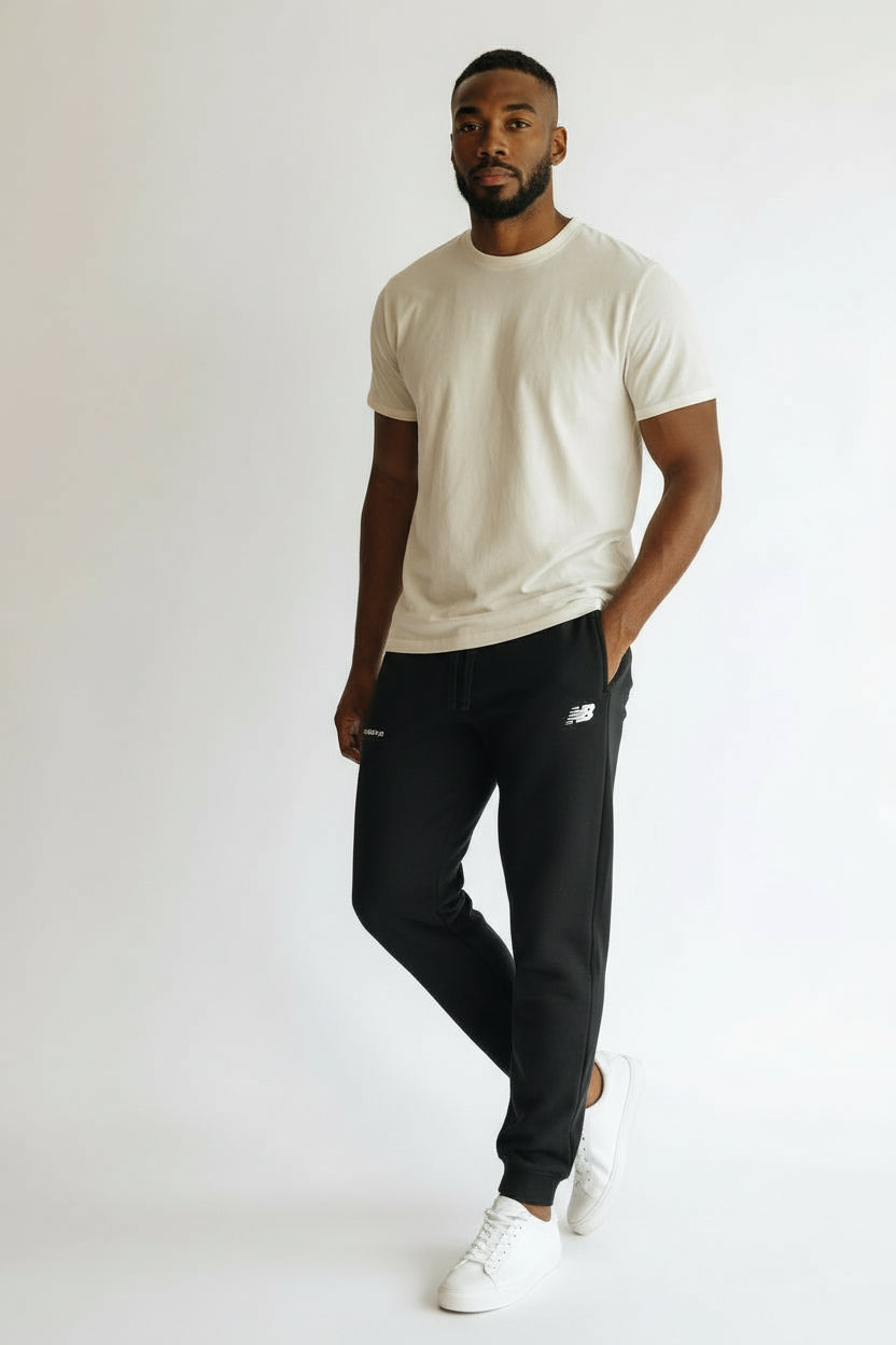 Man wearing a beige t-shirt, black pants, and white sneakers on a plain background. New Balance, Monza Outlet, Monza Fashion, Shein Outlet