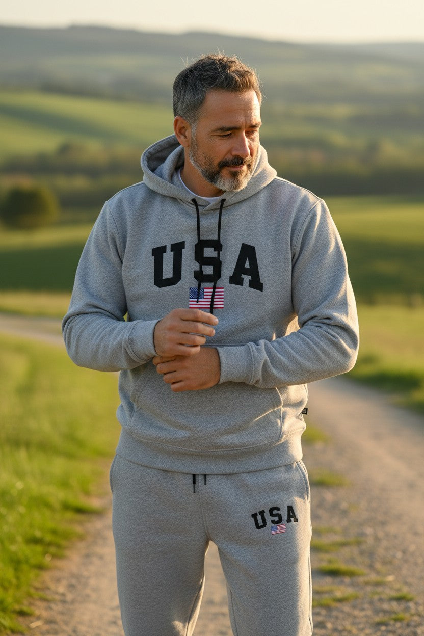 Man wearing a gray USA hoodie and pants standing on a dirt path with green fields in the background. 