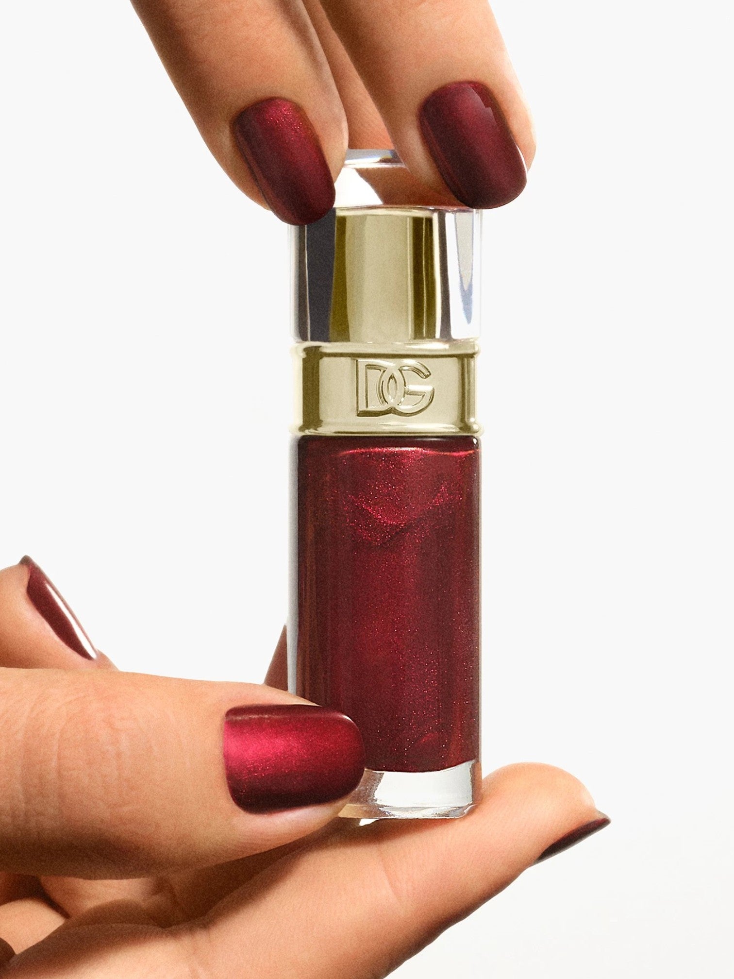 A hand holding a clear glass bottle of metallic deep red nail polish with a shiny gold cap, against a plain white background. Dolce and Gabanna, Monza Outlet, Monza Fashion, Shein Outlet