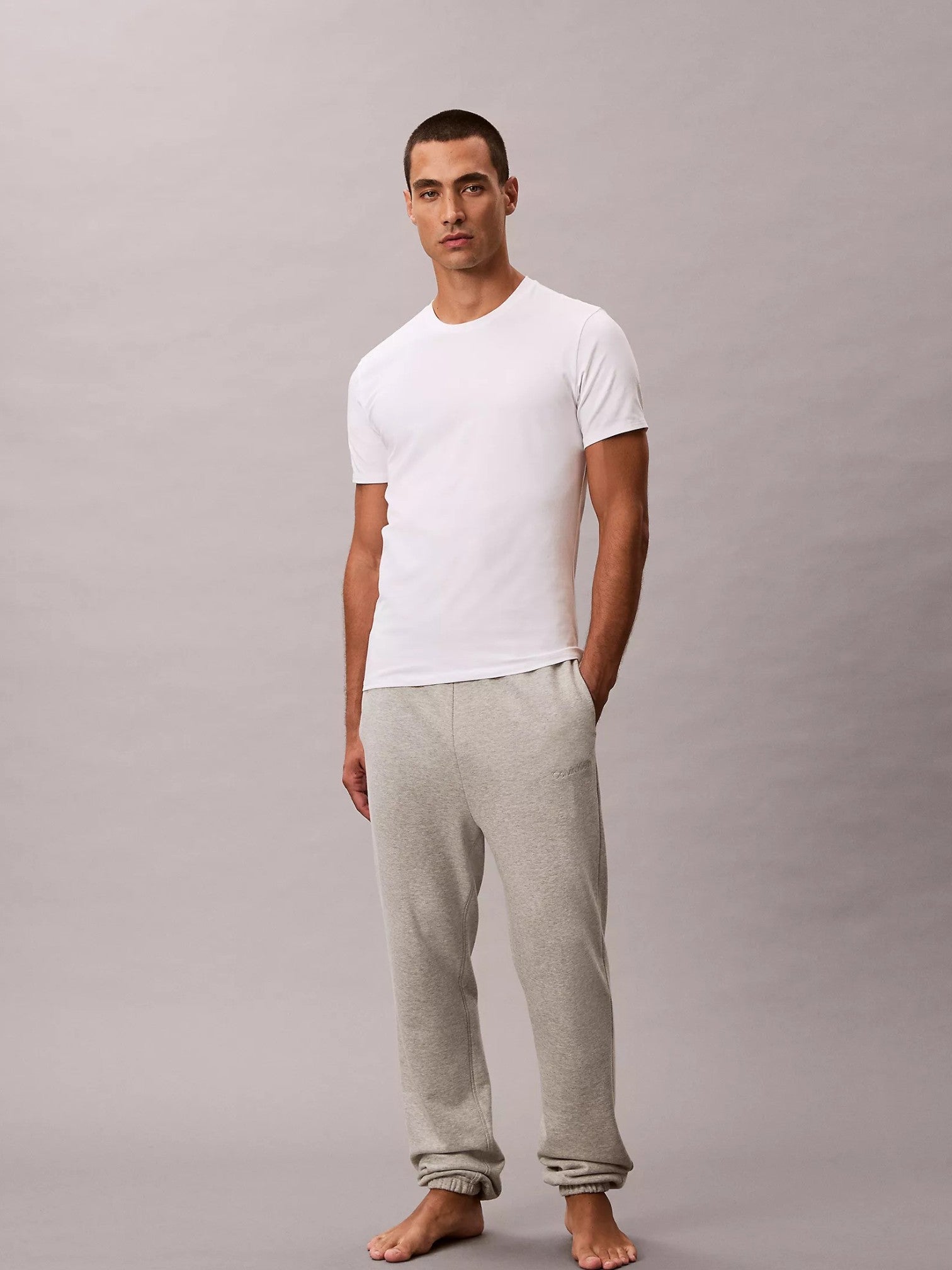 A man standing barefoot against a plain light gray studio background, wearing a plain white short-sleeve crew-neck tshirt and light heather gray jogger sweatpants. Calvin Klein, Monza Outlet, Monza Fashion, Shein Outlet