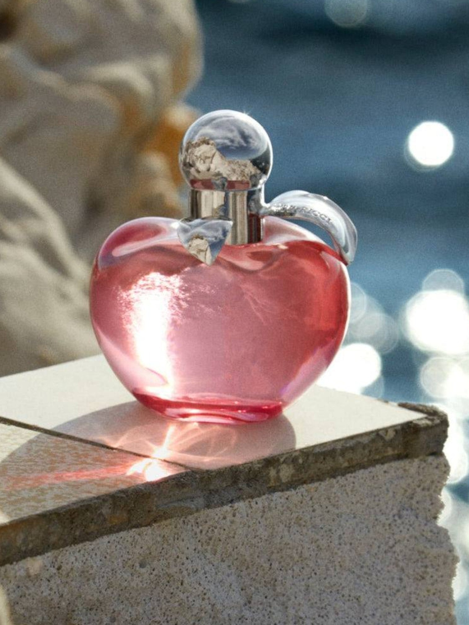 Pink perfume bottle with silver cap on a stone surface by a body of water. Nina Ricci, Monza Outlet, Monza Fashion, Shein Outlet