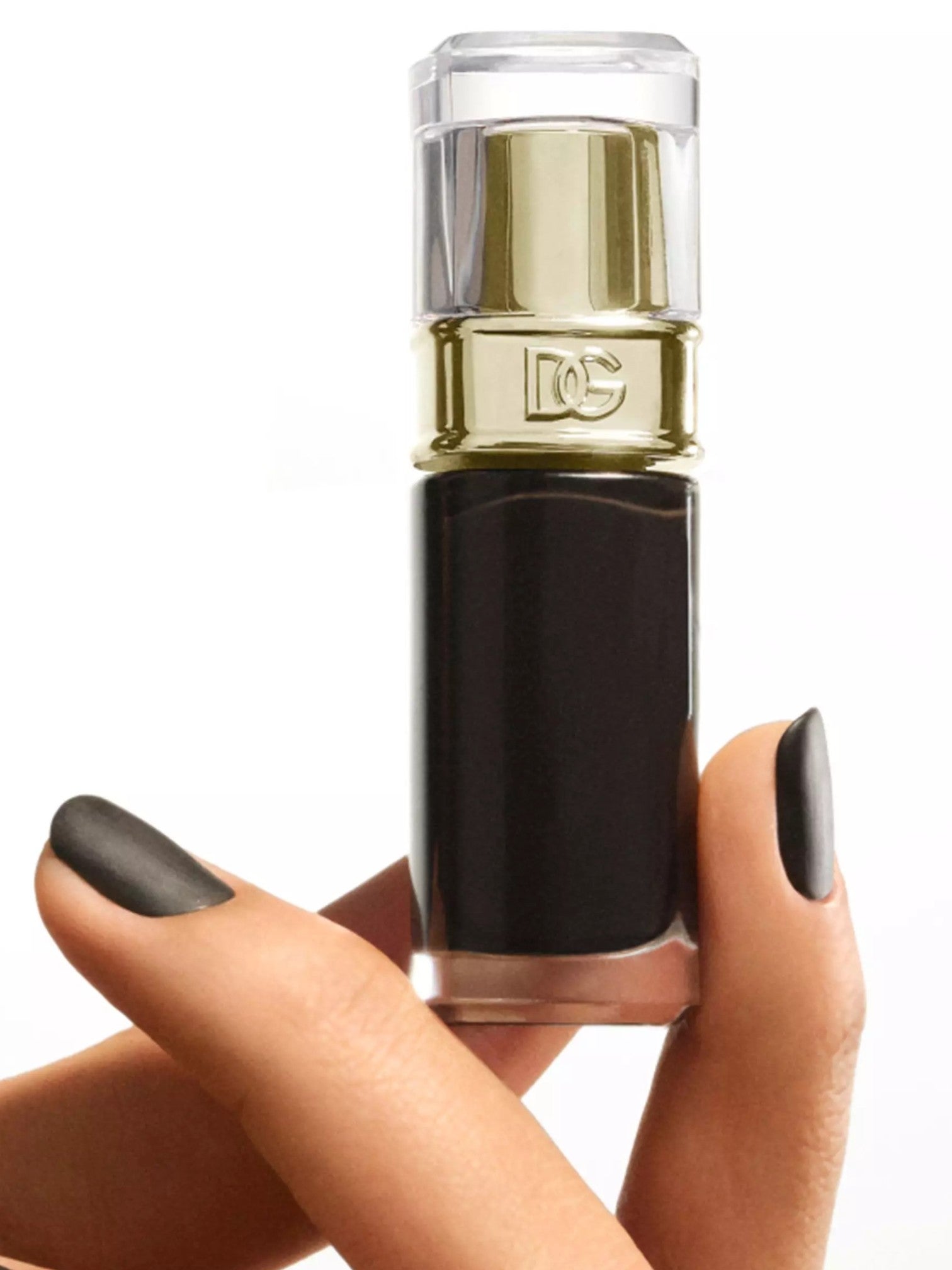 A hand with dark gray painted nails holds a cylindrical cosmetic bottle with a black body, gold metallic collar, and clear cap against a white background. Dolce and Gabanna, Monza Outlet, Monza Fashion, Shein Outlet