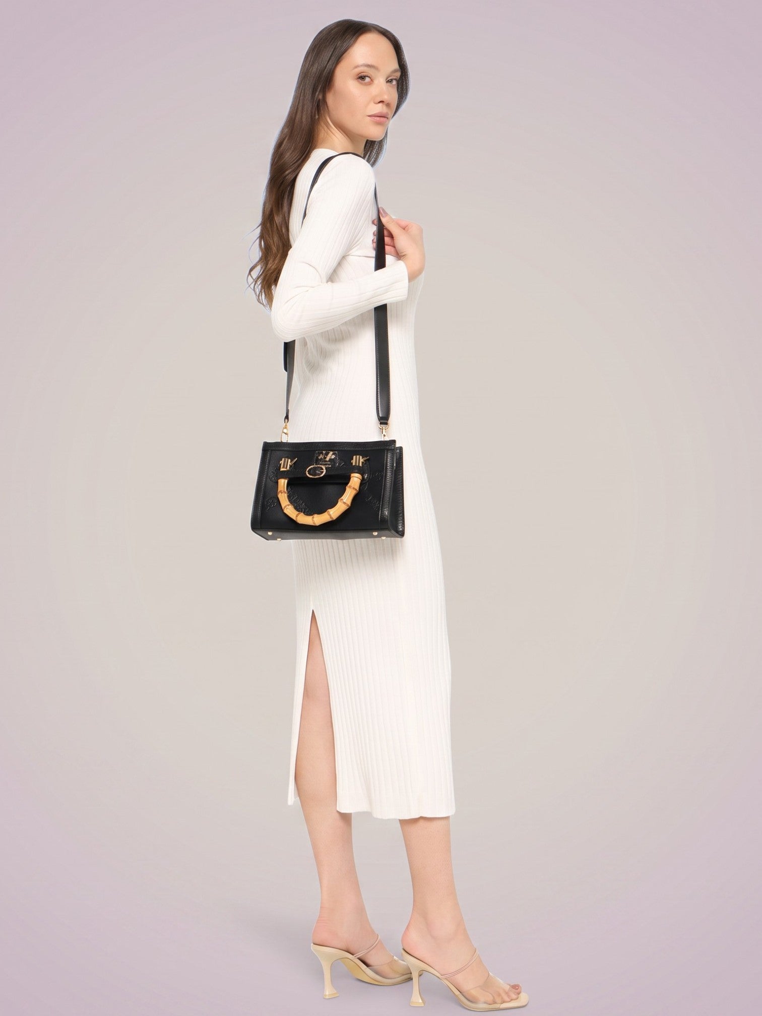 A woman wearing a white ribbed long sleeve midi dress with a side slit, styled with a black shoulder bag featuring a gold tone circular handle and strap, and beige mule heels, against a solid light lavender studio background. 19v69 Italia, Monza Outlet, Monza Fashion, Shein Outlet