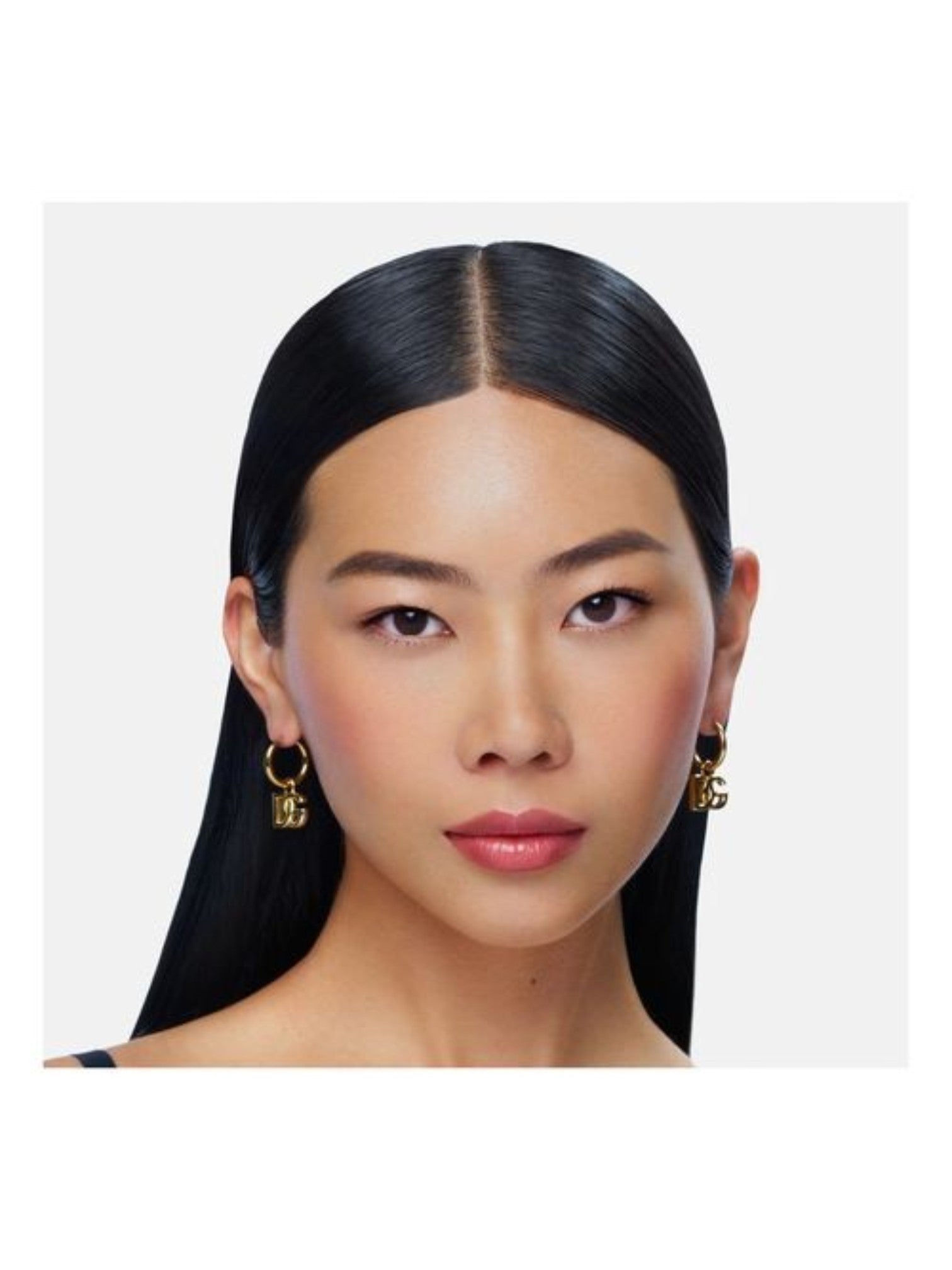 A closeup portrait of a woman with long, straight black hair wearing small gold hoop earrings with dangling charms, set against a light gray background. Dolce & Gabbana, Monza Outlet, Monza Fashion, Shein Outlet
