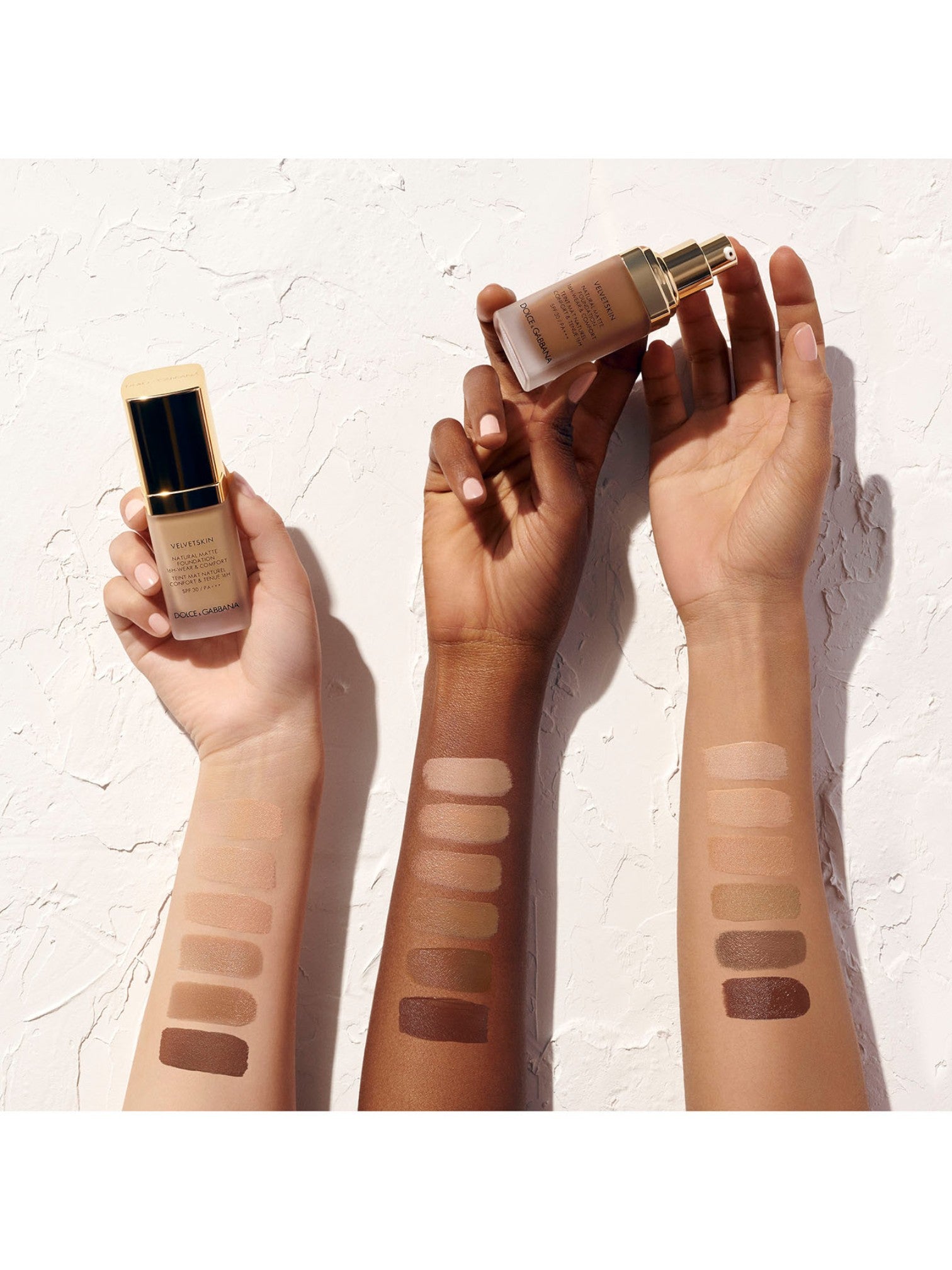 Three hands holding a foundation bottle with swatches on a textured white background Dolce Gabbana Monza Outlet