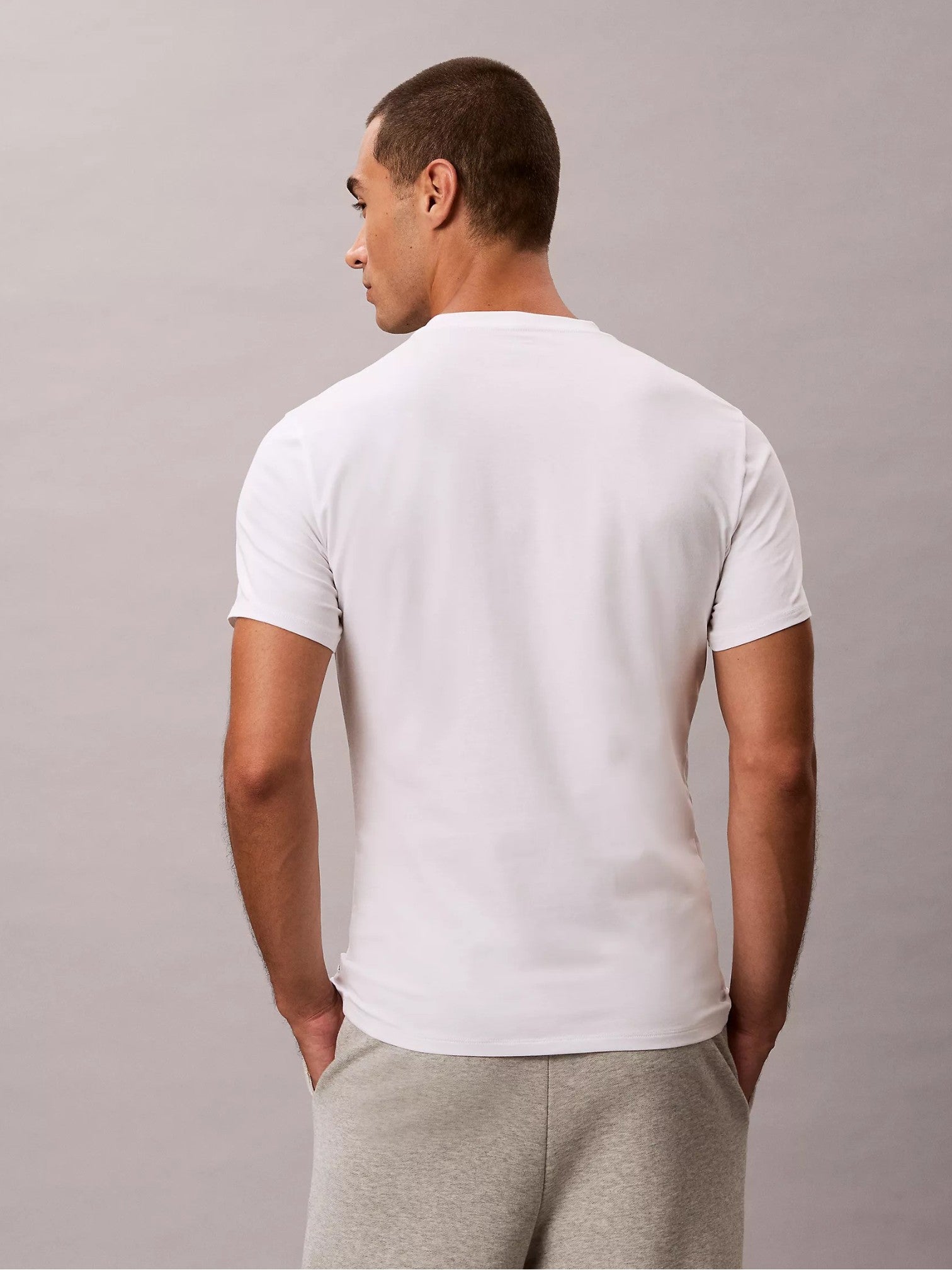 A man viewed from the back wearing a plain white short-sleeve crewneck tshirt and light gray sweatpants, standing against a neutral gray studio background. Calvin Klein, Monza Outlet, Monza Fashion, Shein Outlet