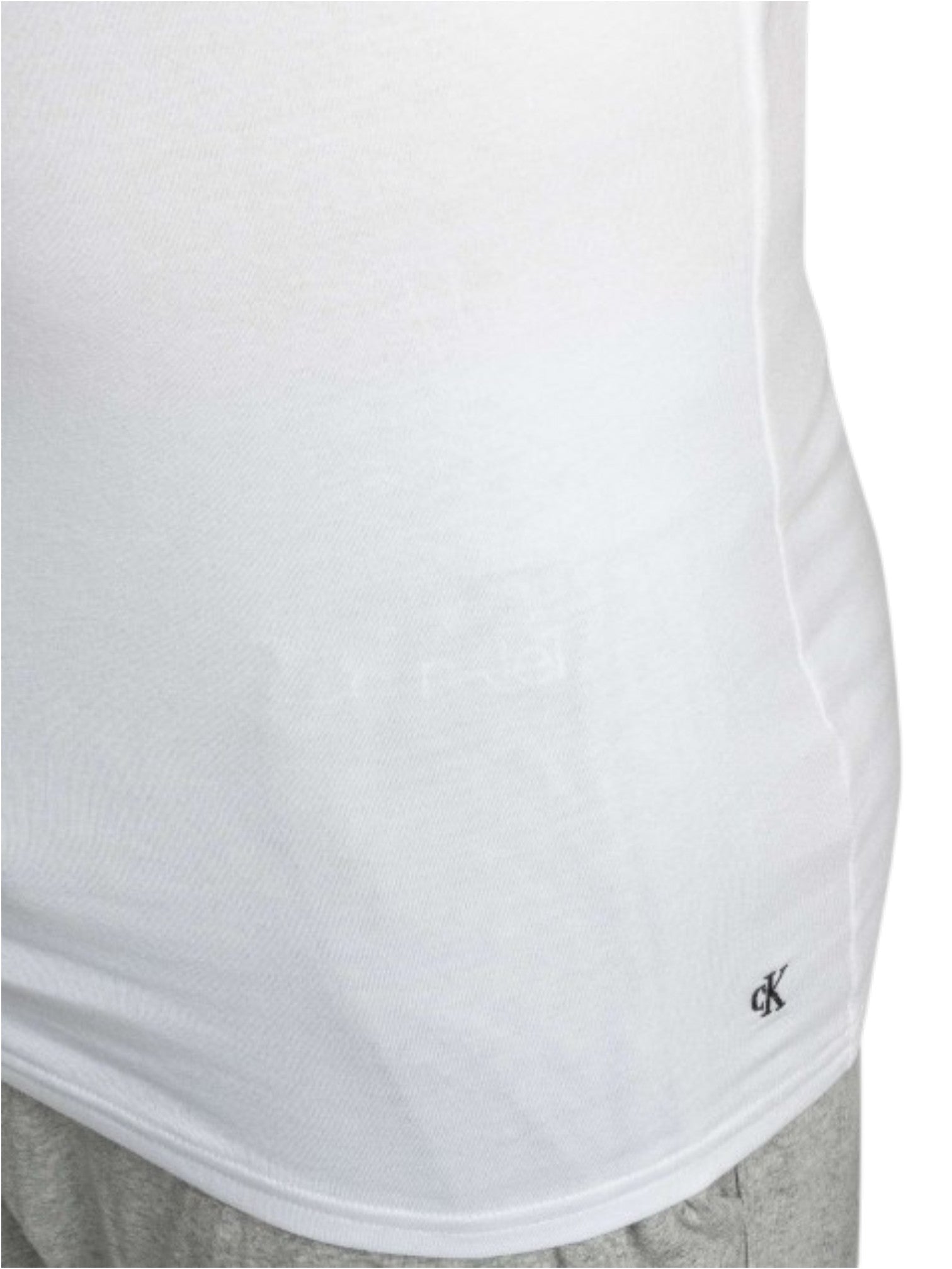 Close-up of a white athletic tank top showing the side seam and a small black Calvin Klein logo near the lower hem, paired with gray shorts. Calvin Klein, Monza Outlet, Monza Fashion, Shein Outlet