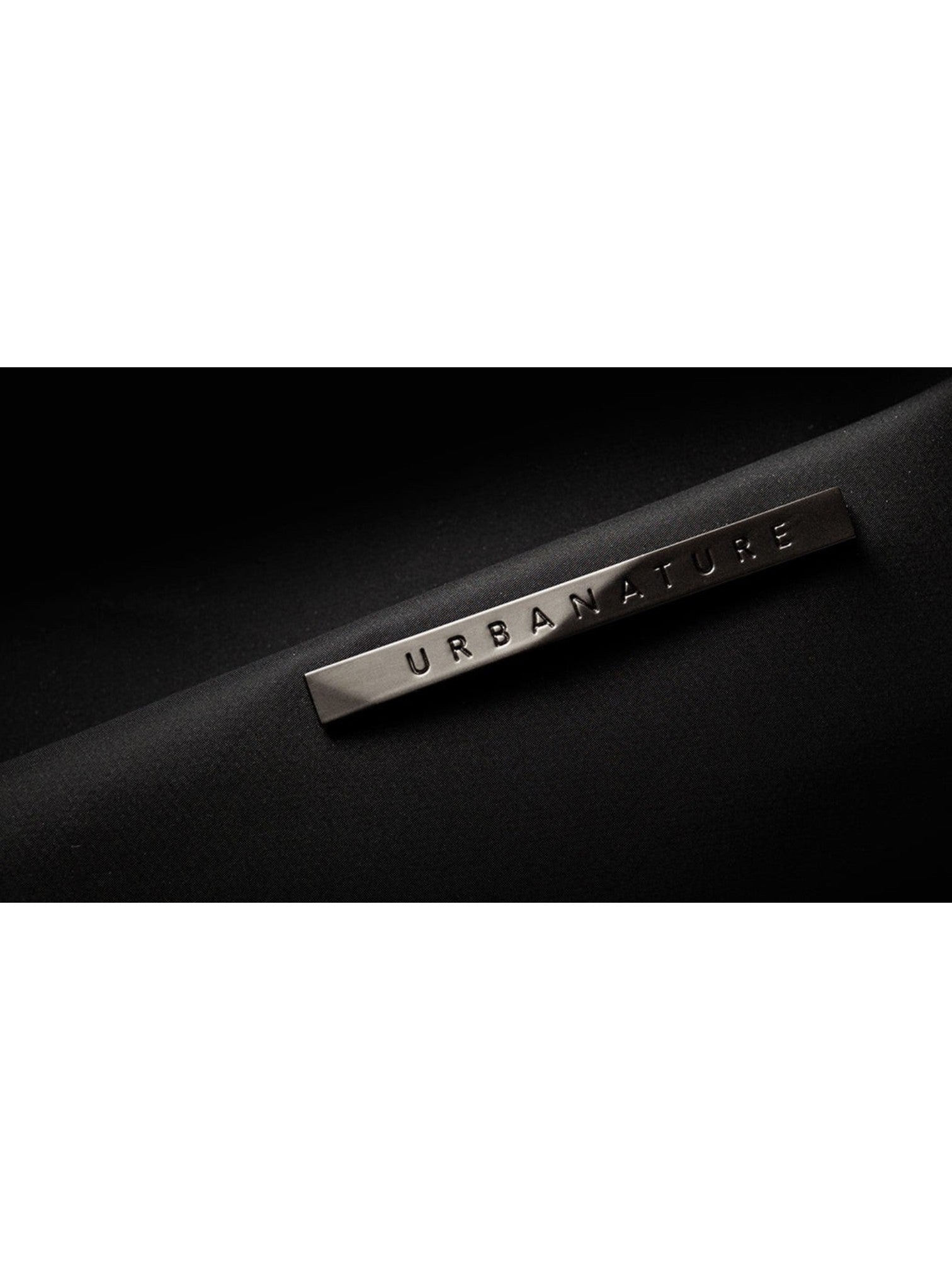 A close-up of a black textured fabric item featuring a rectangular silver metal brand plaque engraved with URBANATURE. NIIDR, Monza Outlet, Monza Fashion, Shein Outlet