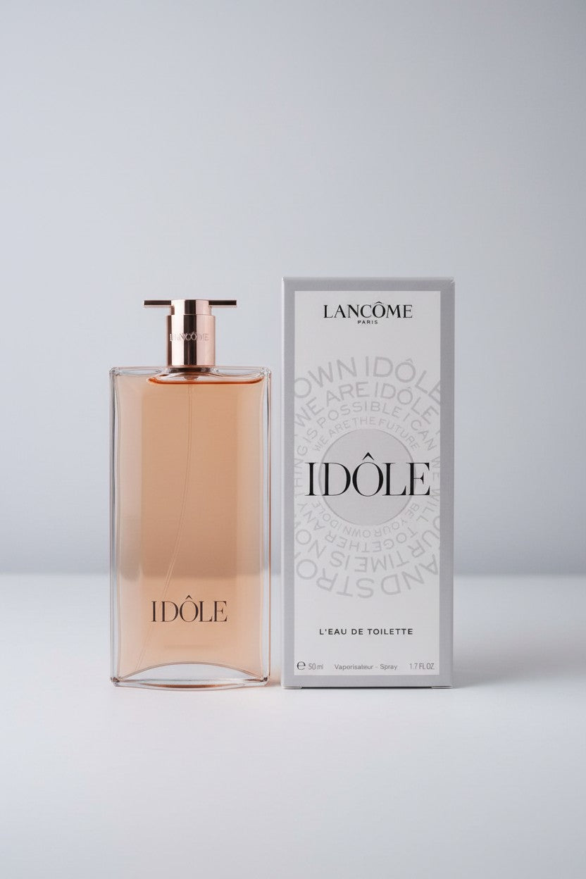 A bottle of Lancôme Idôle Eau de Toilette perfume with peach colored liquid and a rose gold cap, standing next to its white and gray packaging box on a light gray background. Lancome, Monza Outlet, Monza Fashion, Shein Outlet