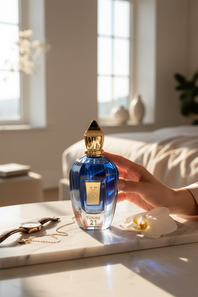 A hand holds a blue glass perfume bottle with a gold cap and a gold label that reads PARNASSI 21 on a marble tray in a bright, sunlit bedroom. Xerjoff, Monza Outlet, Monza Fashion, Shein Outlet