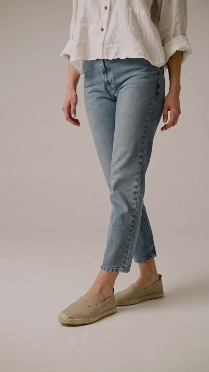 A model wearing light wash blue cropped jeans, a white button-up shirt, and beige slip on shoes, standing against a plain light beige studio background. Tommy Hilfiger, Monza Outlet, Monza Fashion, Shein Outlet