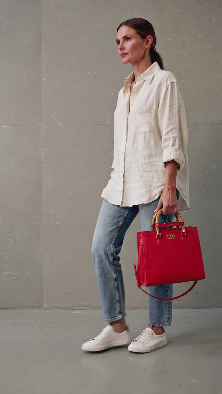 A woman wearing a white linen shirt, blue jeans, and white sneakers holds a structured red handbag with gold tone hardware and bamboo handles against a gray concrete wall. 19v69 Italia, Monza Outlet, Monza Fashion, Shein Outlet
