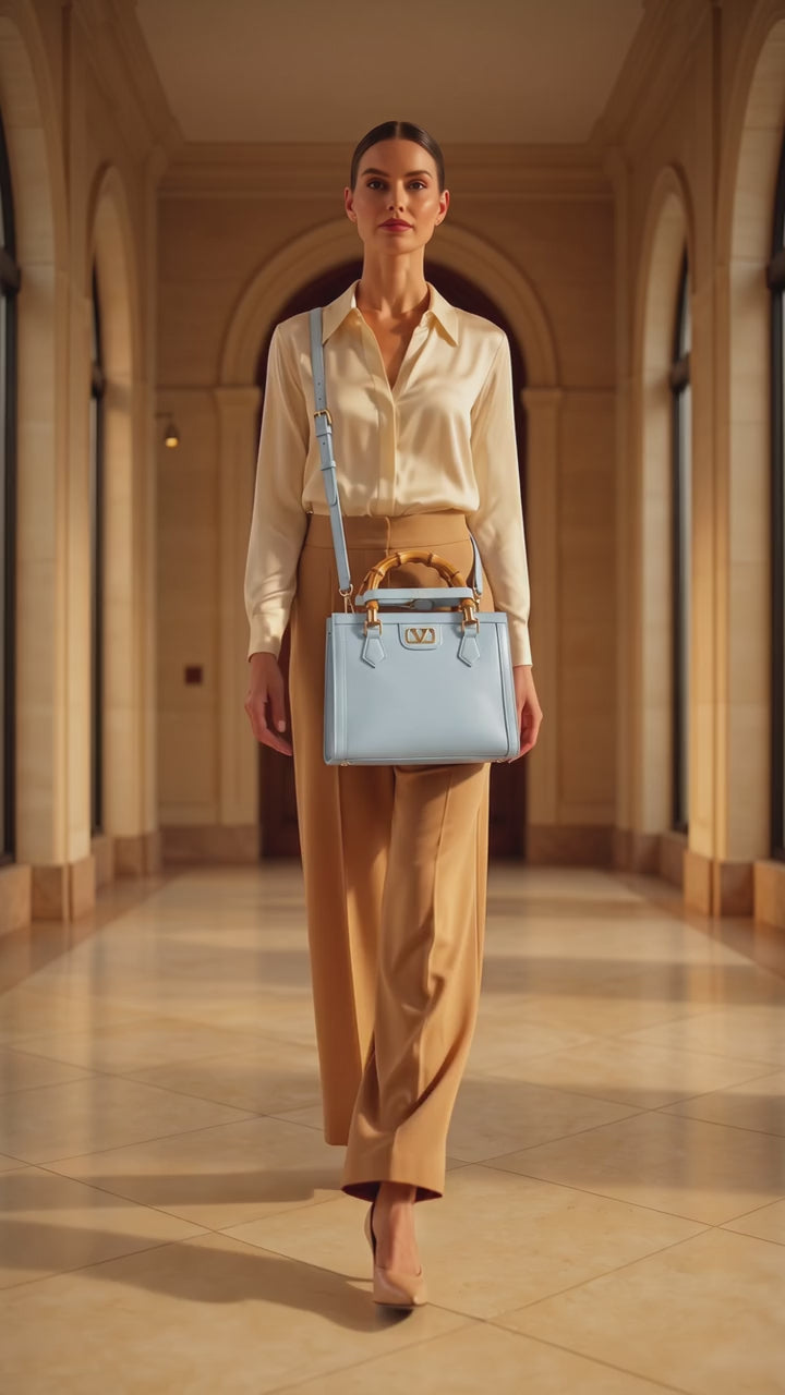A woman walks down a grand, sunlit hallway wearing a cream satin shirt, tan wide leg trousers, and beige heels, while carrying a structured light blue handbag with gold hardware and a bamboo style top handle. 19v69 Italia, Monza Outlet, Monza Fashion, Shein Outlet